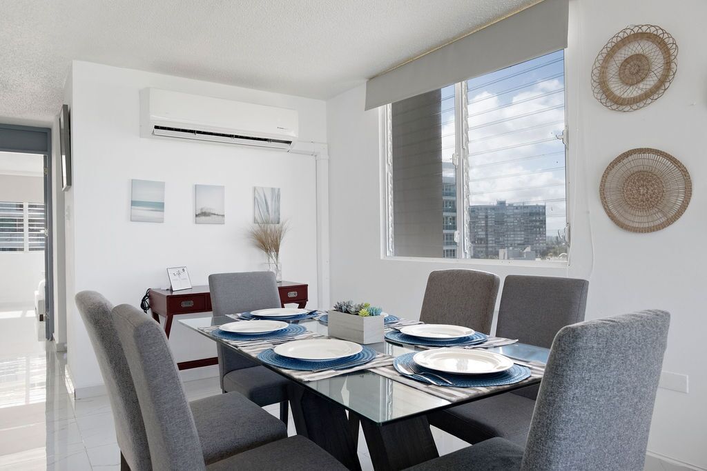 A modern dining room with a glass table, grey upholstered chairs, and a city view through large windows.