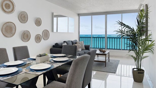 A modern living room with a dining area, large windows overlooking the sea, and a balcony with a railing. - Flamenco Beach 2BDR Oceanfr Pool Parking (Carolina)