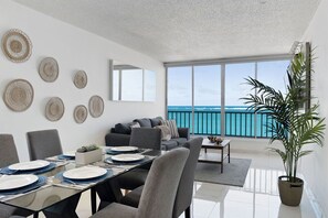 A modern living room with a dining area, large windows overlooking the sea, and a balcony with a railing. - Flamenco Beach 2BDR Oceanfr Pool Parking (Carolina)