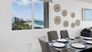 A dining area with a table set for four, a cityscape view through a large window, and decorative woven wall art. - Flamenco Beach 2BDR Oceanfr Pool Parking (Carolina)