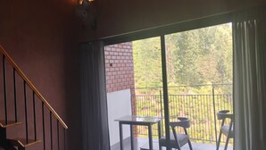 A room with a staircase, a table, and chairs on a balcony with a view of a brick wall and trees.