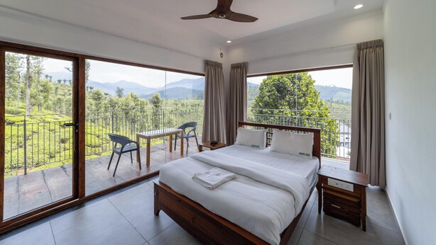 A bedroom with a large bed, a ceiling fan, a wooden nightstand, and a view of a lush landscape through large windows.