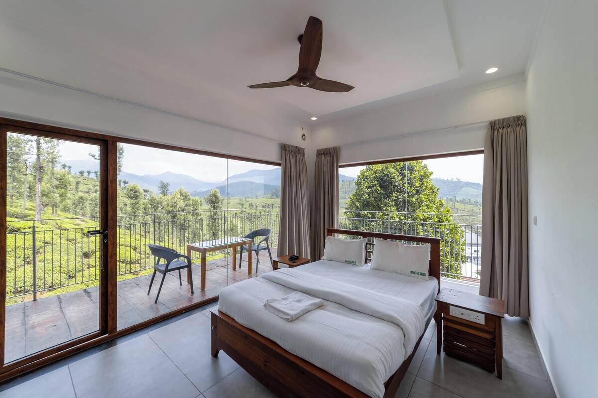 A bedroom with a large bed, a ceiling fan, a wooden nightstand, and a view of a lush landscape through large windows.
