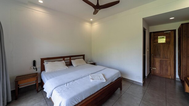 A bedroom with a single bed, a wooden nightstand, a ceiling fan, and a wooden door. - WINTEL WILD (Valparai)