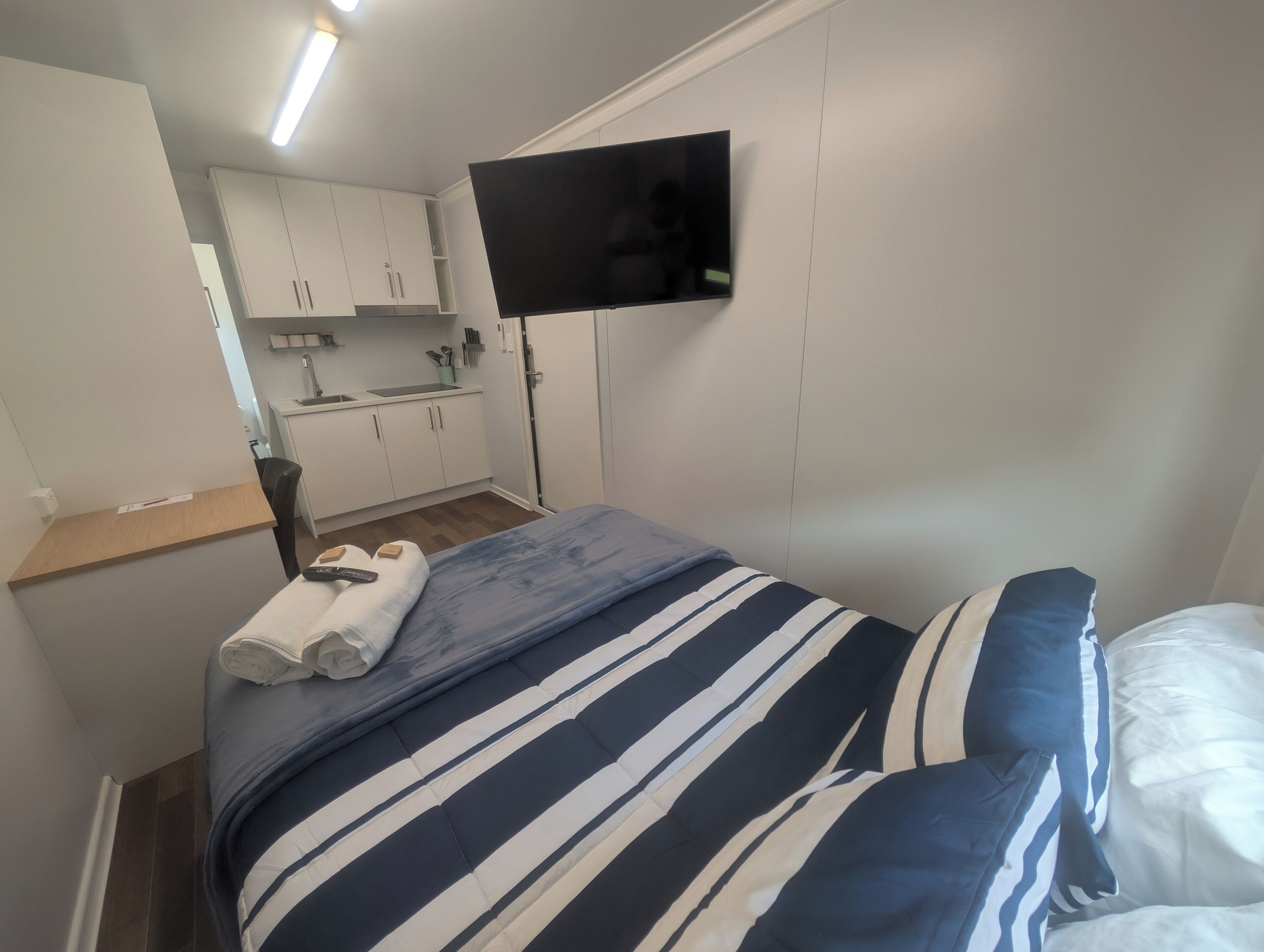 A bedroom with a bed, a TV mounted on the wall, a small kitchenette, and a bathroom.