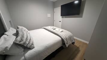 Een netjes opgemaakt bed met witte lakens en een zachte deken, een flatscreen televisie aan de muur bevestigd en een houten vloer.