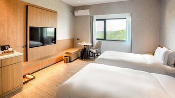 Habitación de hotel con dos camas, un televisor montado en un panel de madera, una mesita con una silla y una ventana con vistas a zonas verdes.