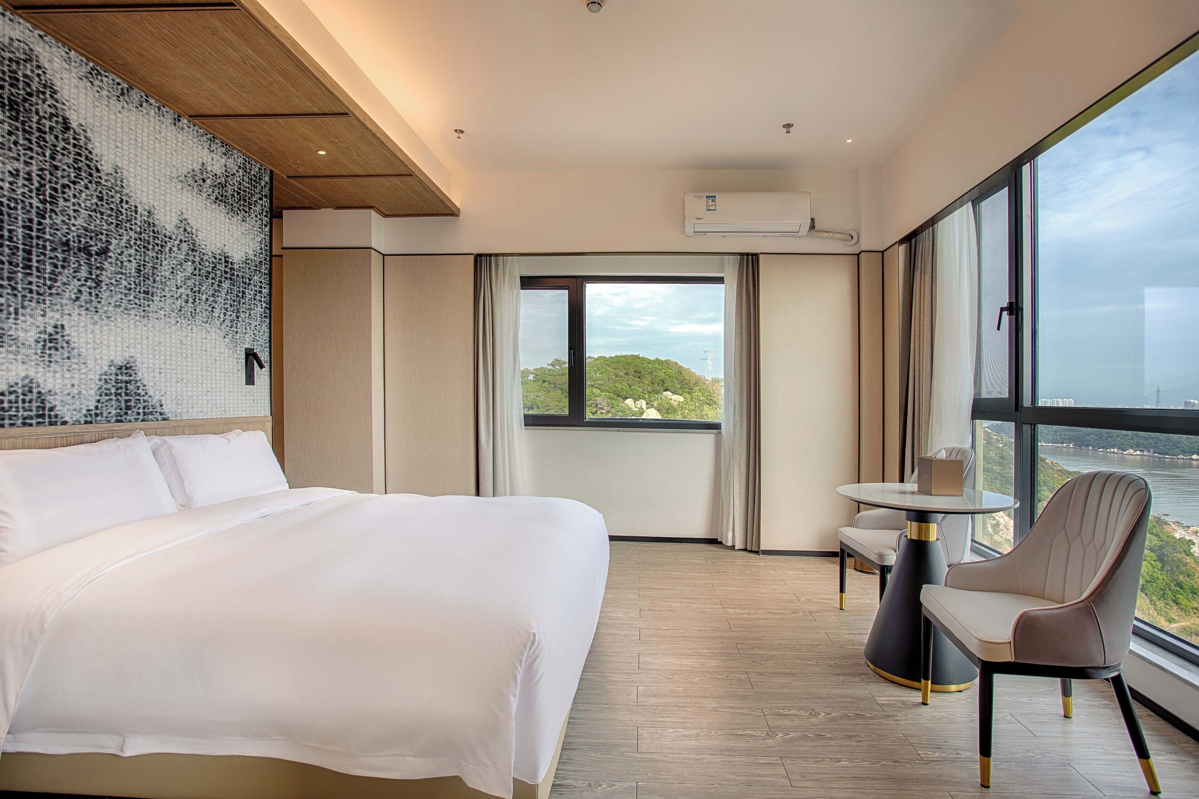A hotel room with a large bed, a small table, a chair, and a view of a body of water through large windows.