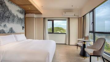 A hotel room with a large bed, a small table, a chair, and a view of a body of water through large windows.
