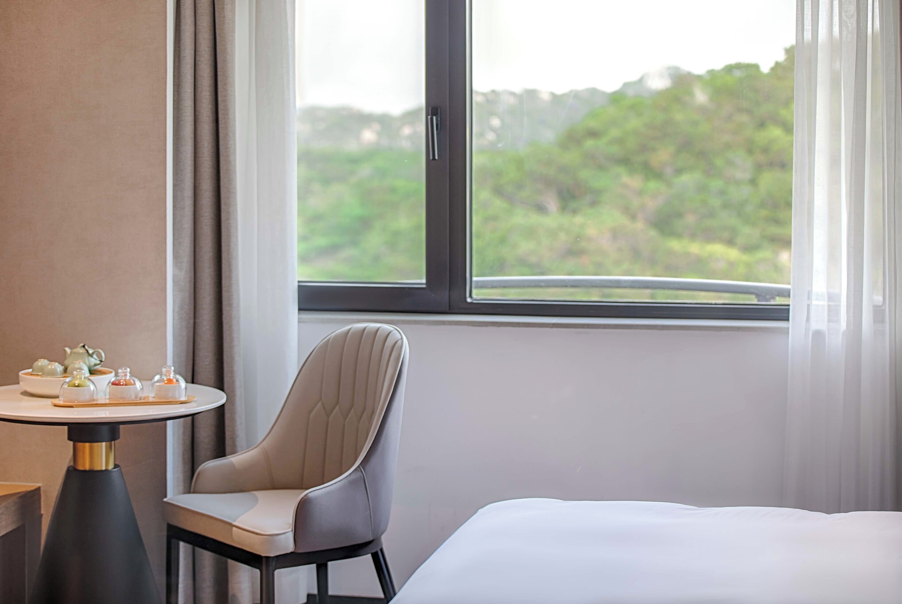 A hotel room with a bed, a chair, a table with tea sets, and a window with a view of greenery.