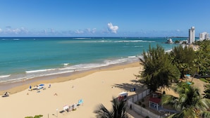 Una playa con agua azul cristalina, una orilla arenosa y algunos sombrillas. Se observan edificios al fondo y palmeras a lo largo de la playa.