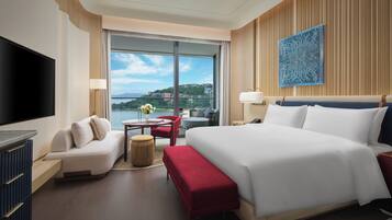 A modern hotel room with a large bed, a TV, a sofa, a small table, and a view of the city.