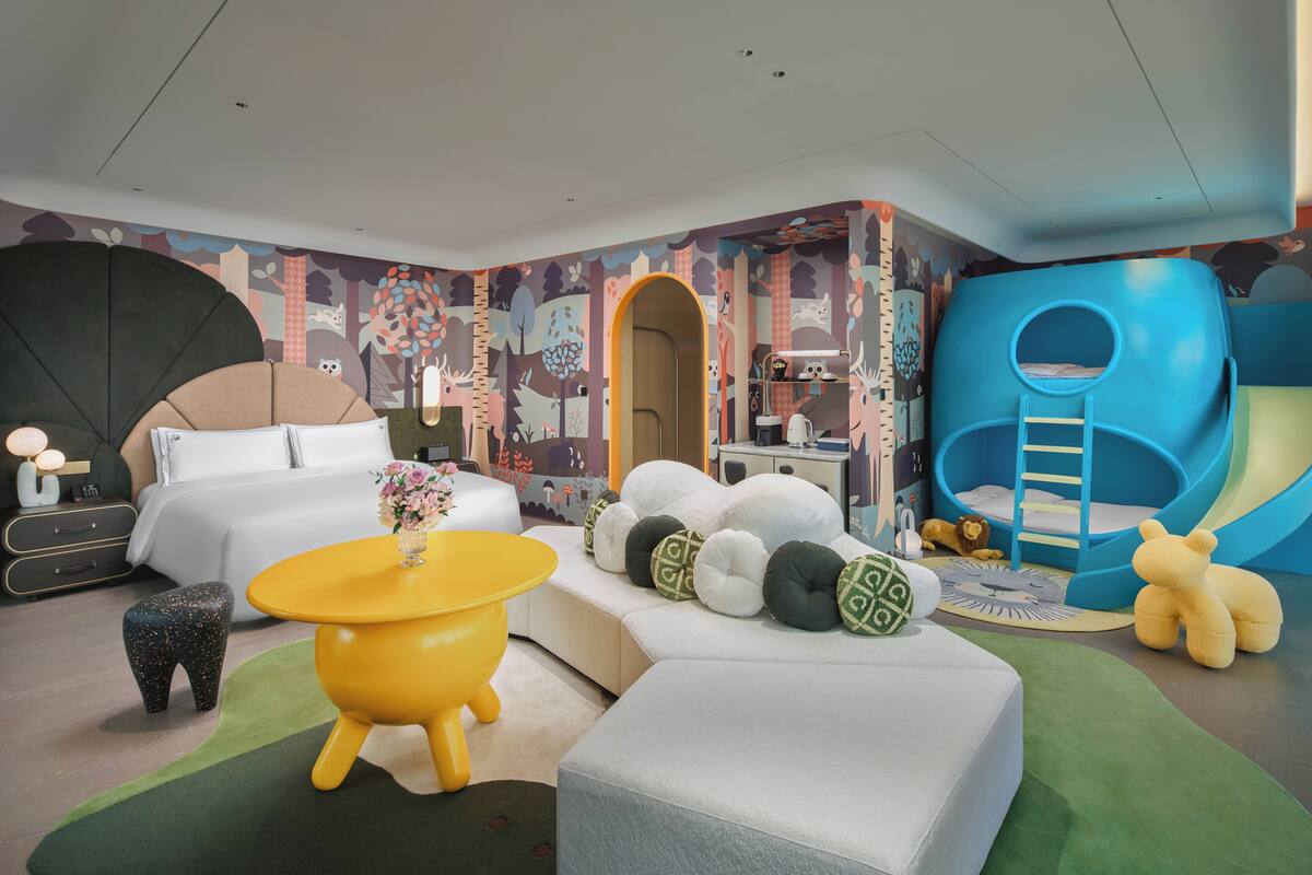 Et moderne hotelværelse med seng, sofa, et gult bord og et legeområde med en rutchebane.