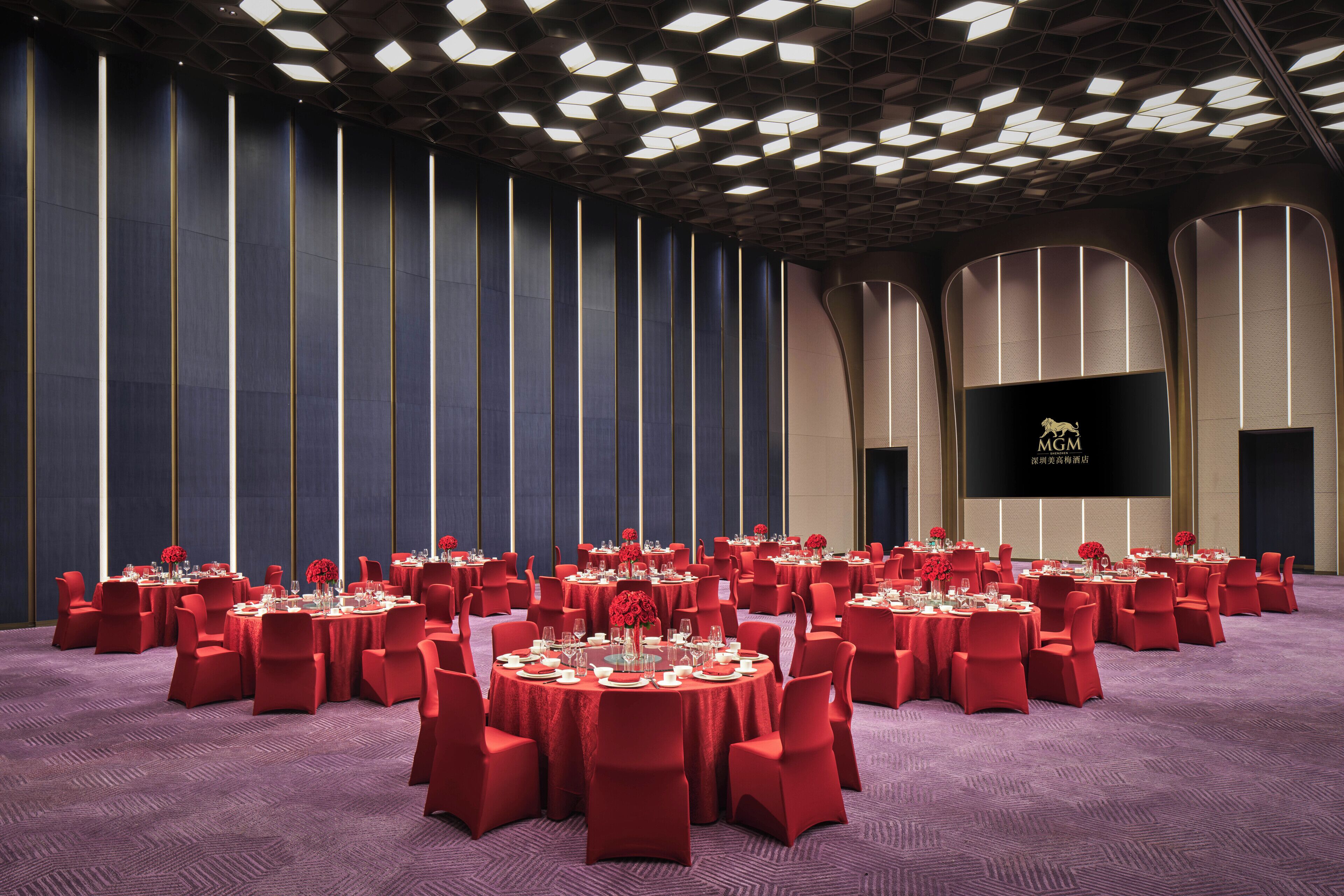 Une salle de conférence avec des tables rondes, des chaises rouges et un grand écran affichant du texte.