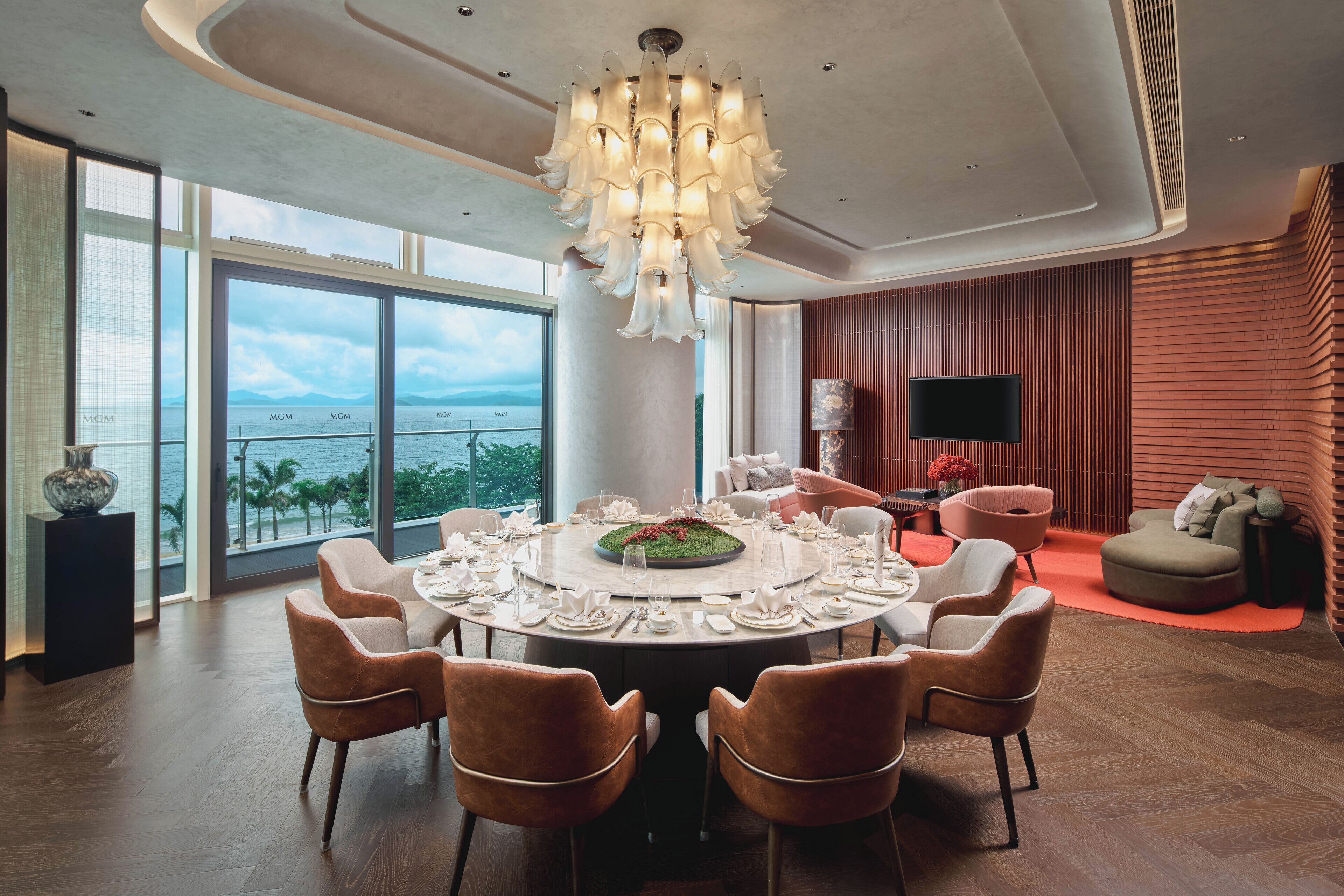 A modern dining area with a round table set for six, a large chandelier, and a view of the ocean through large windows.
