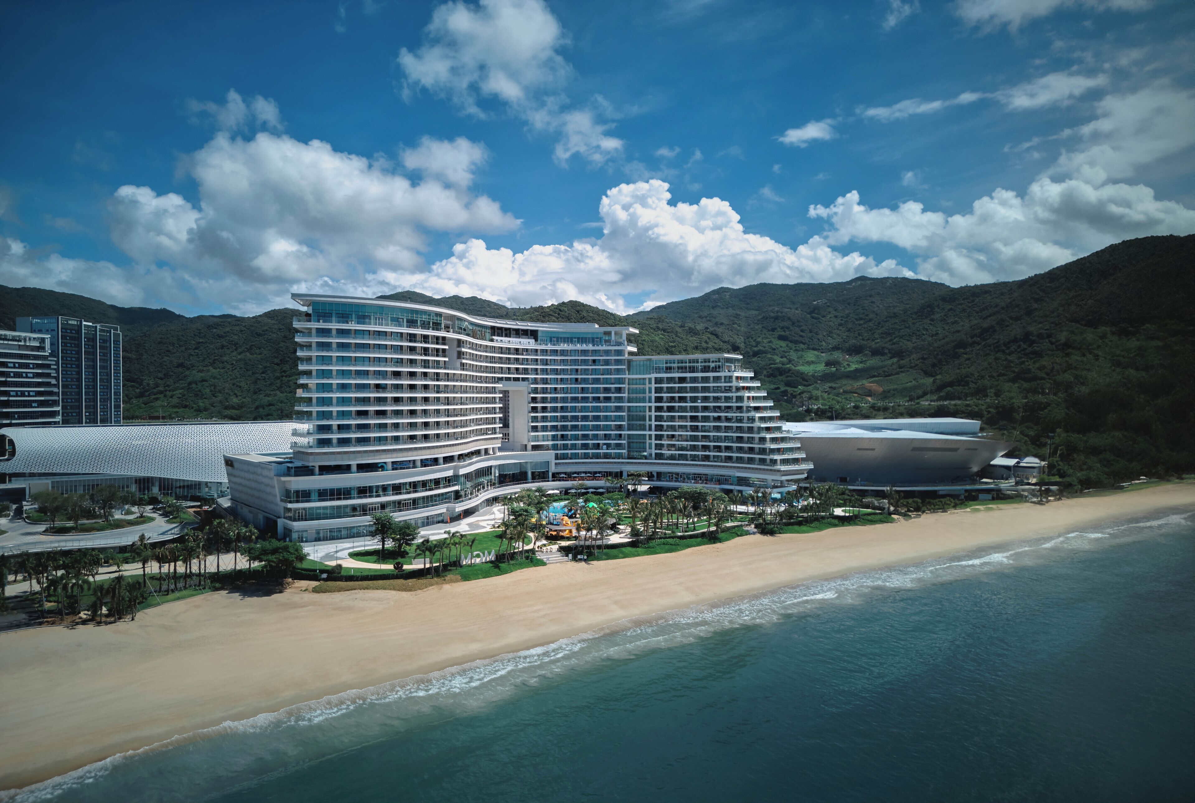 Um hotel grande e moderno, com vários andares e um design curvo, localizado na praia, com vista para o oceano e montanhas ao fundo.
