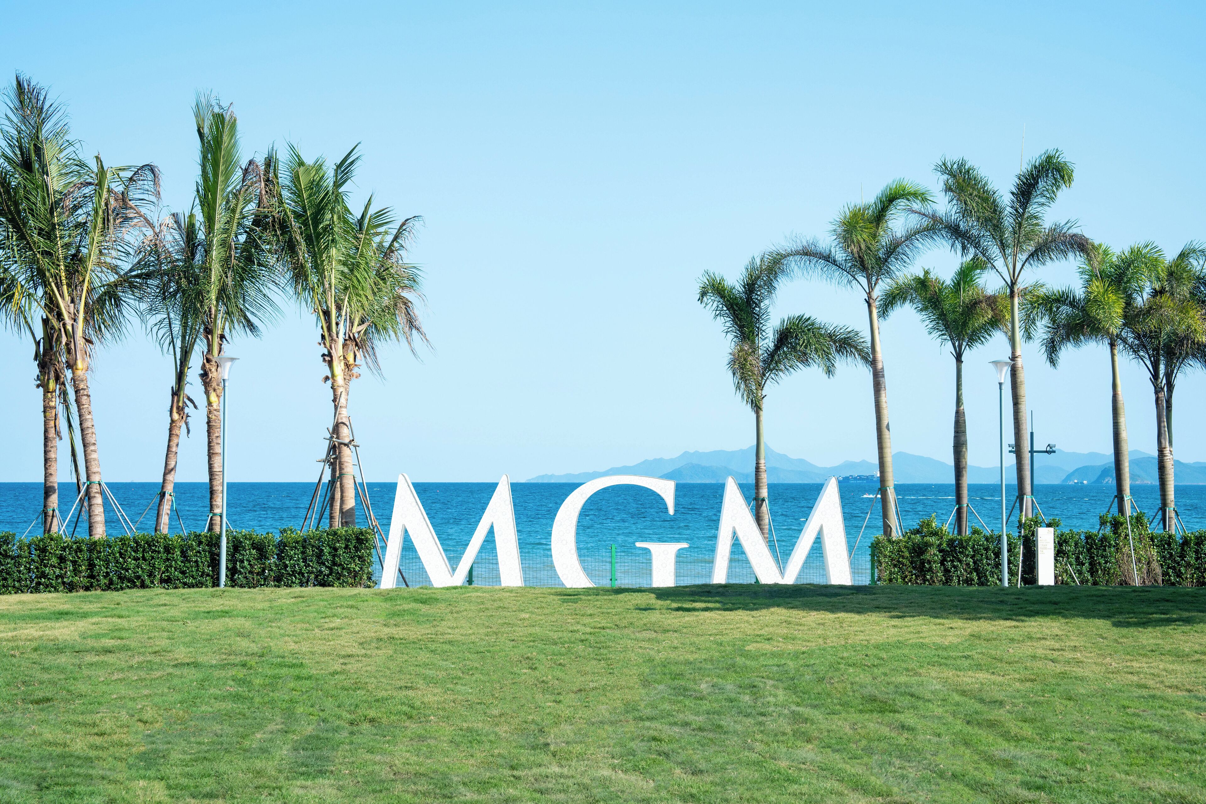 A grassy area with palm trees and a large 'MGM' sign in the foreground, with a clear view of the ocean and mountains in the background.