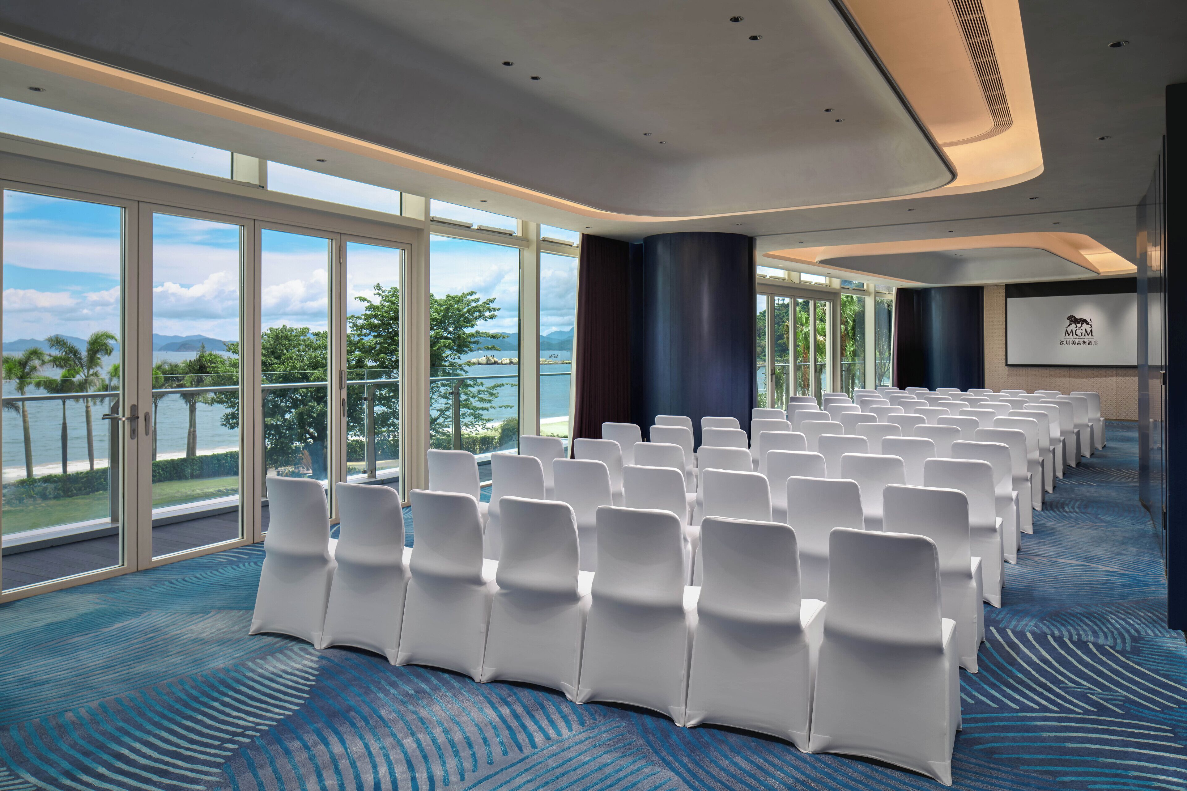 Un salón de conferencias con filas de sillas blancas, ventanas amplias con vista al mar y una pantalla mostrando el logotipo de MGM.