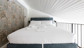 Ein ordentlich bezogenes Bett mit weißen Leinen und gefalteten Handtüchern, ein dunkles Kopfbrett und eine Wand mit floralem Wandpapier.