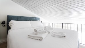Un lit bien fait, avec du linge de lit blanc et des serviettes pliées, une tête de lit bleue recouverte de tissu et un plafond blanc orné de lignes horizontales.