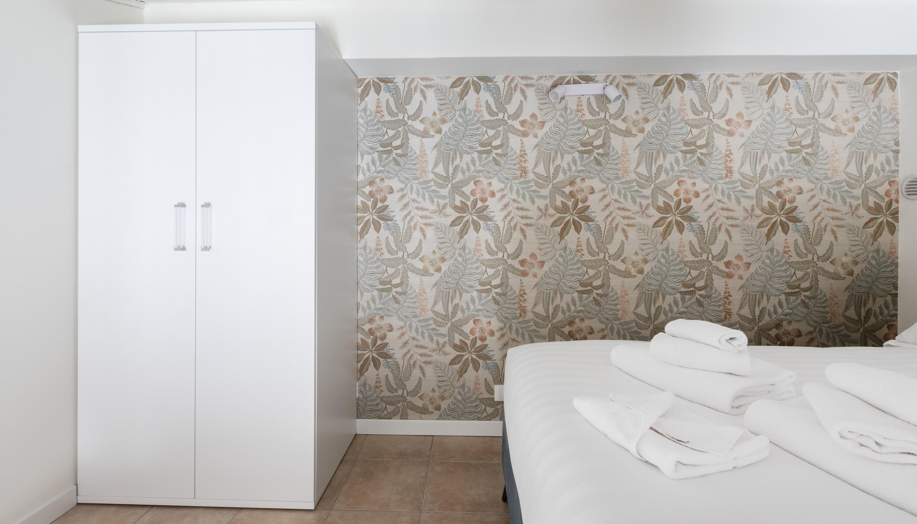 A bedroom with a white wardrobe, floral wallpaper, and neatly folded towels on the bed.