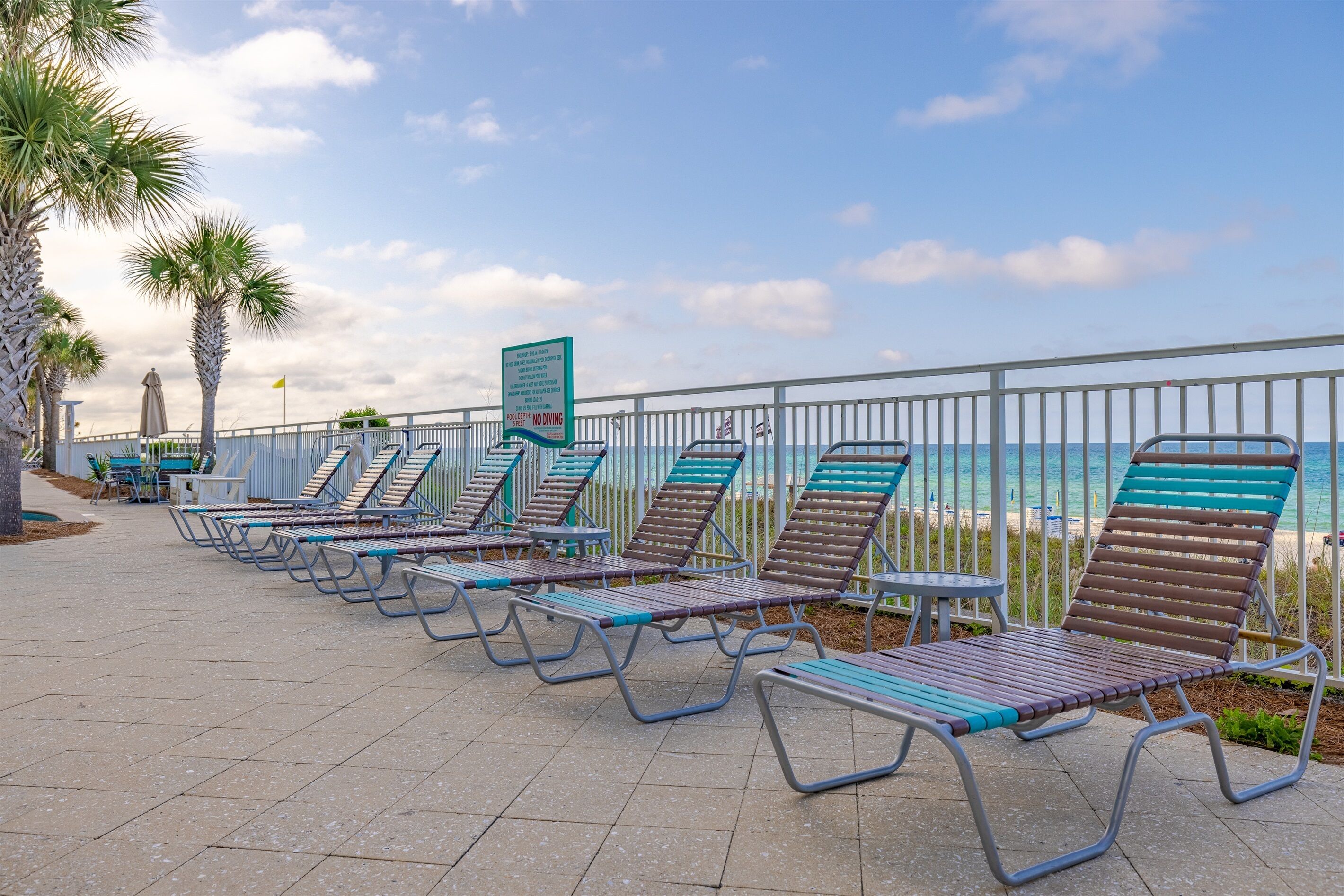 Een rij ligbedden langs een strandboulevard met een metalen hekwerk en palmbomen.