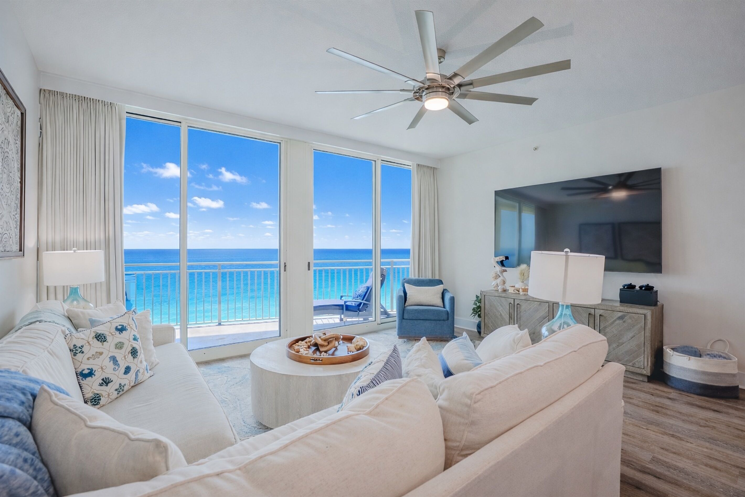 A spacious living room with a large window overlooking the ocean, a ceiling fan, a flat-screen TV, and a comfortable seating area.