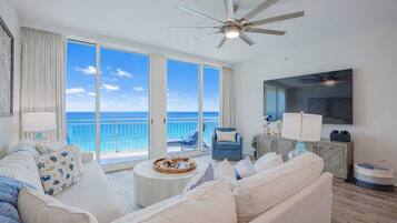 A spacious living room with a large window overlooking the ocean, a ceiling fan, a flat-screen TV, and a comfortable seating area.