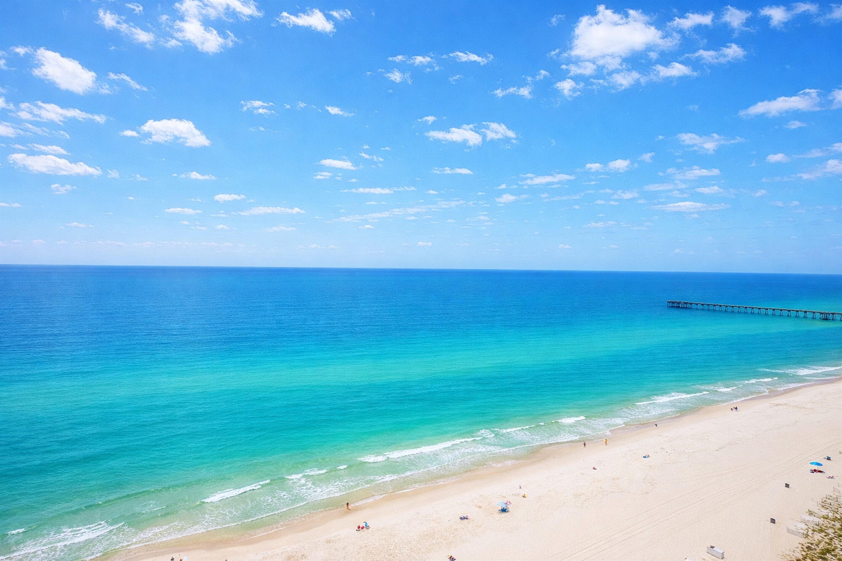 Een strand met helder turquoise water, een pier die in zee uitstelt, en een zandstrand met mensen en parasols.