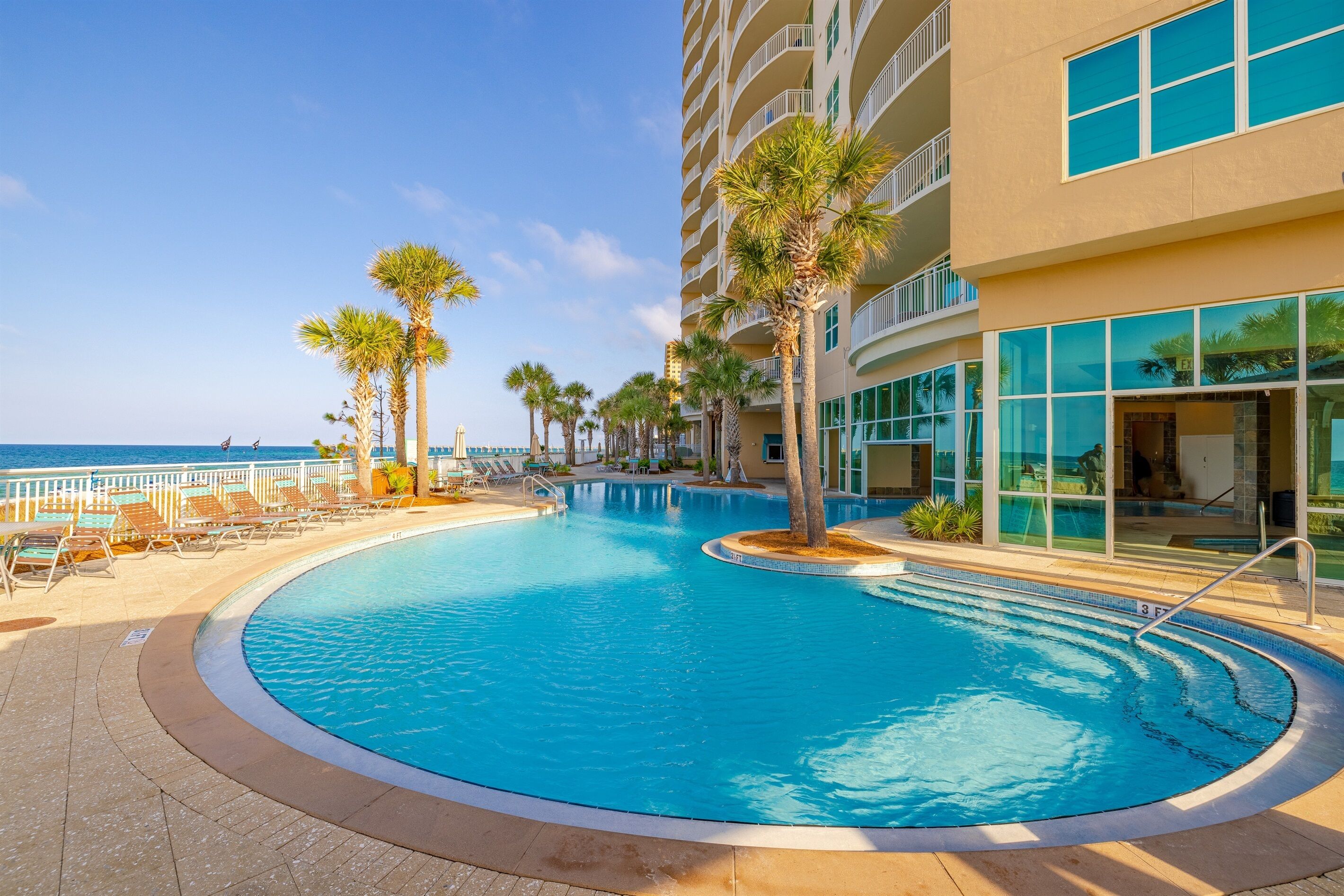Zona de piscina con sillones reclinables y palmeras, rodeada por un edificio con grandes ventanales y balcones.