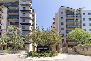 Un edificio residenziale a più piani con balconi, un'area verde alberata e un cartello che indica servizi come un ristorante con cortile e reception.
