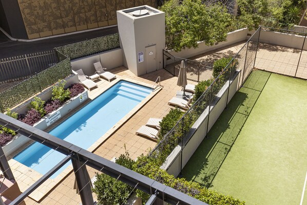 A rooftop swimming pool with lounge chairs, a small building, and a tennis court.