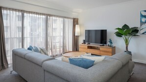A modern living room with a grey sofa, a wooden coffee table, a TV on a wooden stand, and a large window with sheer curtains. - Mounts Bay Stay Two Bedroom Apt.   Near Kings Park - Free Parking - Wi-Fi (Perth)