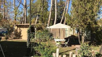 A rustic outdoor seating area with a wooden table and chairs, surrounded by trees and a small wooden structure.