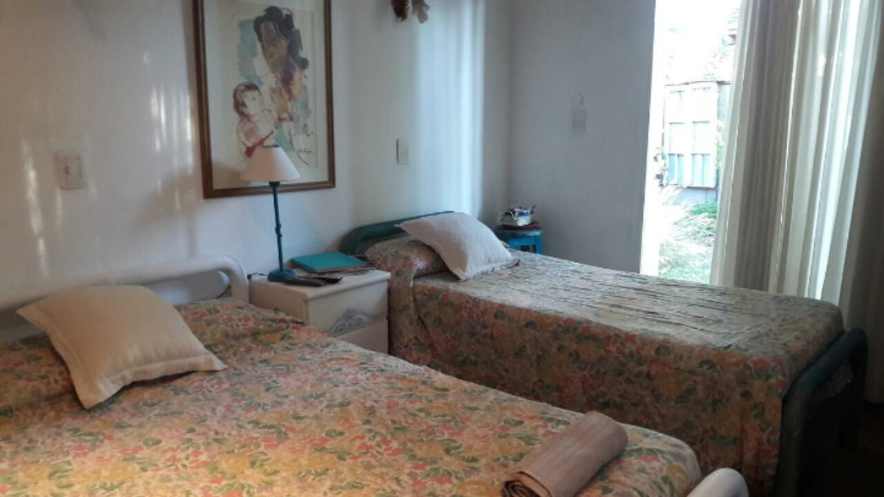 A room with two beds, a nightstand, a lamp, and a window with a view of greenery outside.