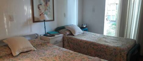 A room with two beds, a nightstand, a lamp, and a window with a view of greenery outside.