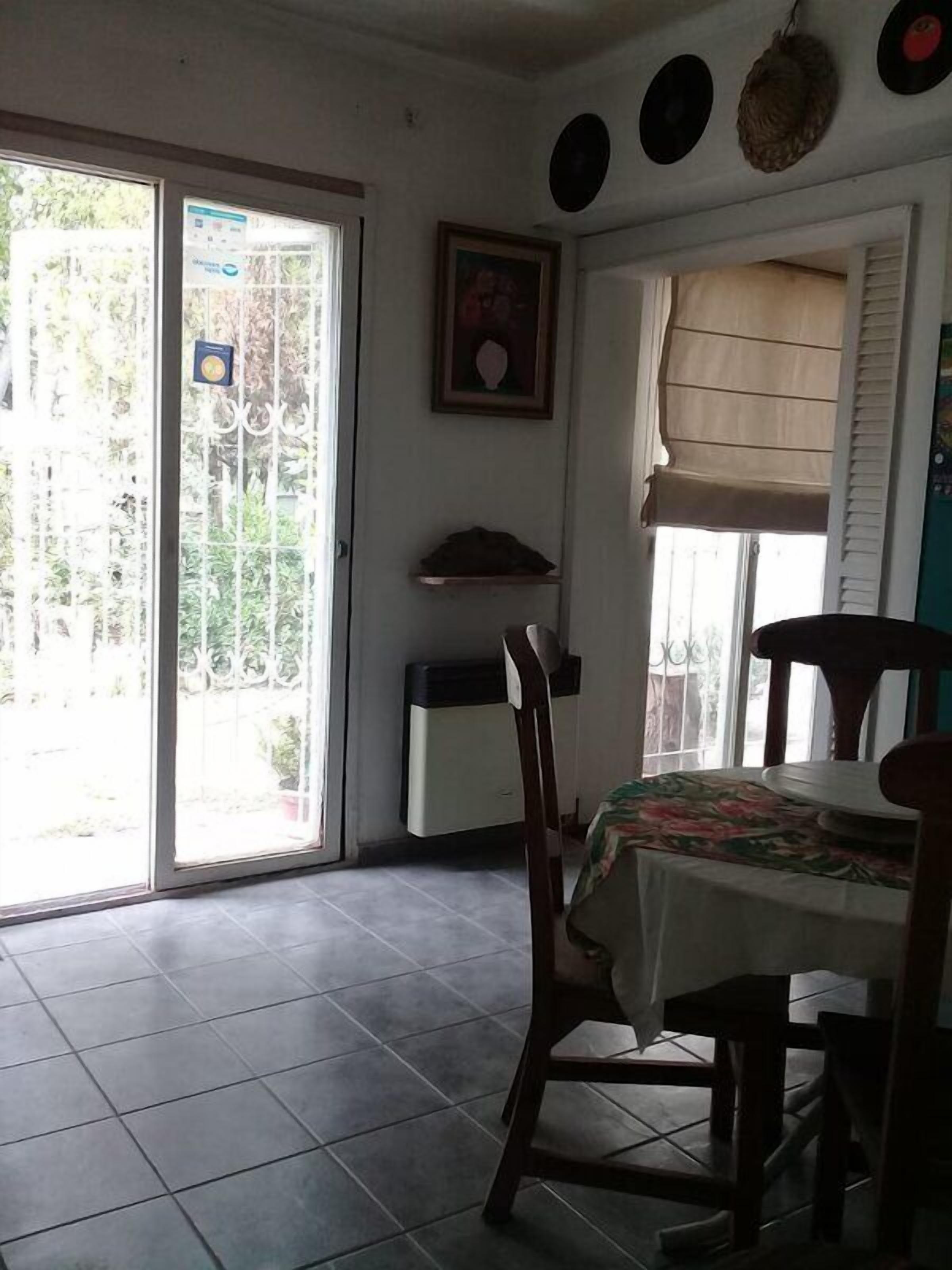 Uma sala de jantar com mesa e cadeiras, uma janela com persianas, uma porta com vista para o verde, e uma parede com um quadro emoldurado e uma prateleira com um objeto preto.