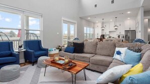 Ein geräumiges Wohnzimmer mit grauem Sofa, blauen Sesseln und einem Holztisch. Im Hintergrund ist eine Küche mit weißen Schränken und Edelstahlgeräten zu sehen.