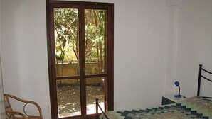 A bedroom with a bed, a chair, a desk with a lamp, and a sliding glass door leading to a garden. - Apartment Type A in Costa Saracena - Castelluccio (Augusta)