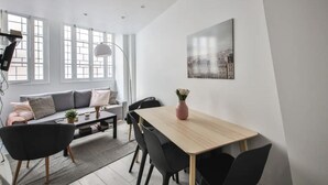 A modern living room with a sofa, a dining table, and chairs. There is a large window with blinds, a floor lamp, and a cityscape painting on the wall. - The Little Rousseau - Cosy & Calm byMon Petit Faré (Paris)