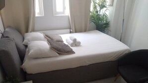 A neatly made bed with pillows and a blanket, a grey sofa, a black chair, a small table with a plant and remotes, and a window with curtains. - The Little Rousseau - Cosy & Calm byMon Petit Faré (Paris)