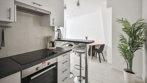 A modern kitchen with a black countertop, stainless steel appliances, and a coffee maker. There is a dining area with a table and chairs, and a potted plant. - The Little Rousseau - Cosy & Calm byMon Petit Faré (Paris)