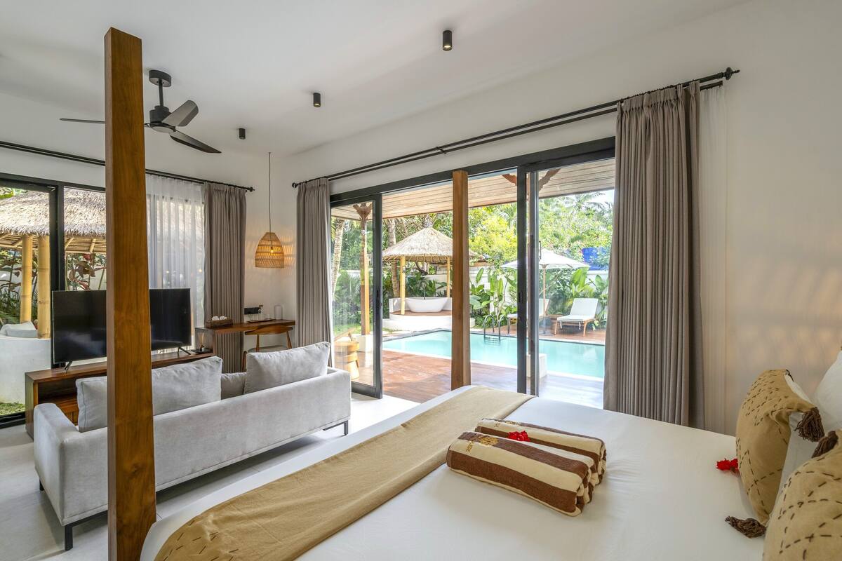 Quarto de hotel moderno com uma cama grande, um sofá, uma TV e vista para uma piscina ao ar livre.