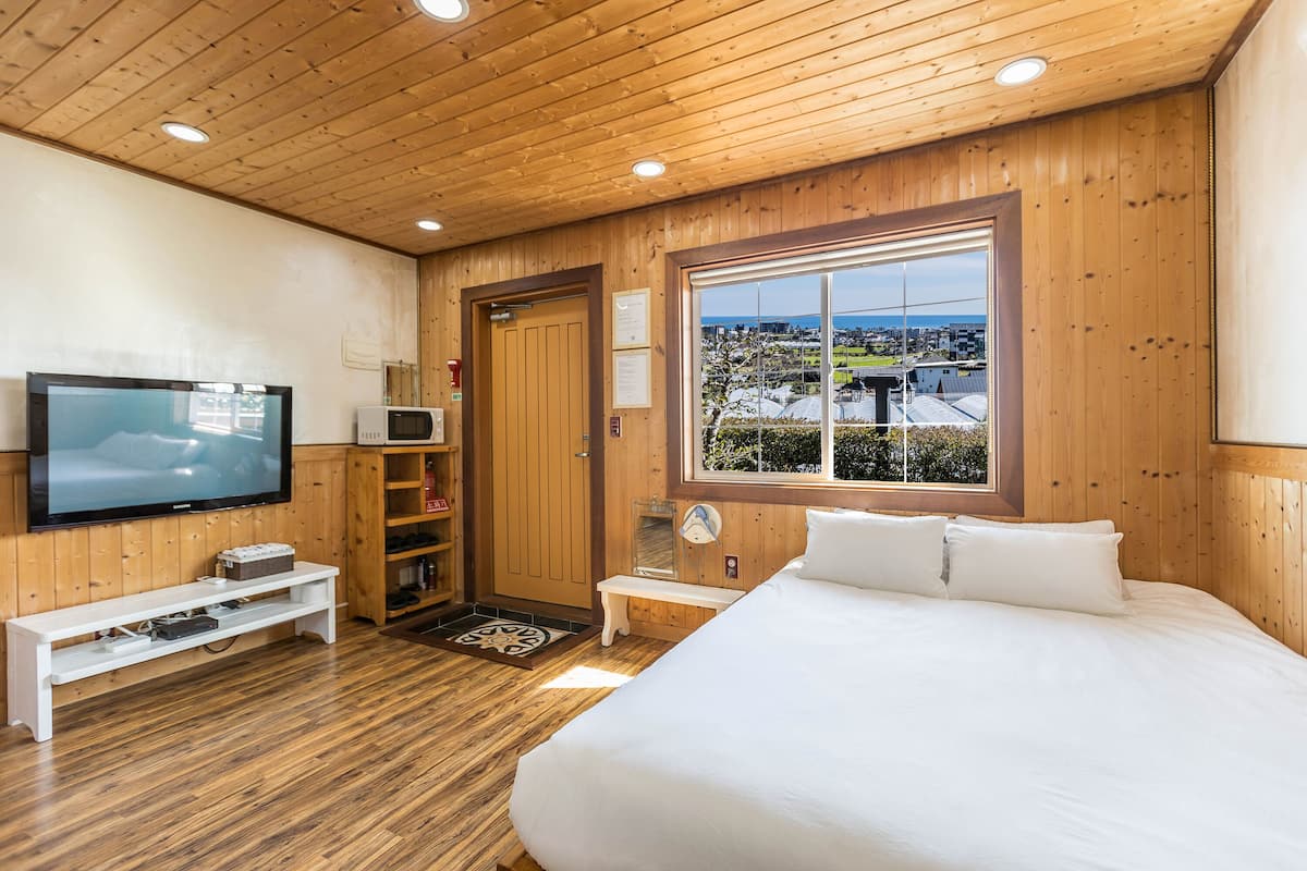 A wooden-paneled room with a bed, a TV, a microwave, and a window with a view of a cityscape.