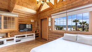 A wooden cabin bedroom with a bed, TV, and large windows overlooking a scenic view. - Habada Pension (Seogwipo)