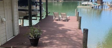 A wooden dock leading to a house with a covered porch, two lounge chairs, and a potted plant.