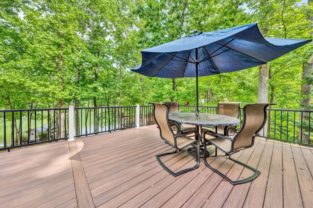 A wooden deck with a table and chairs under a large umbrella, surrounded by a metal railing and lush greenery.