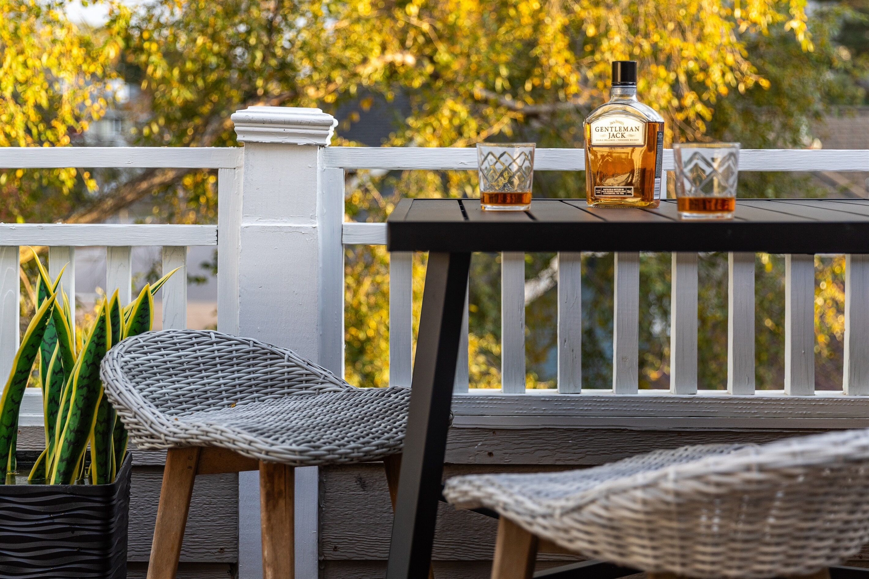 Una terraza de madera con barandilla blanca, una silla de mimbre y una botella de whisky Gentleman Jack sobre una mesita.