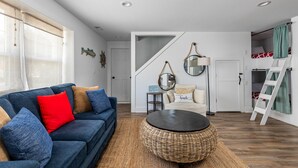 Una sala de estar con un sofá azul, una mesa de centro redonda de mimbre y una escalera que lleva a un dormitorio tipo loft.