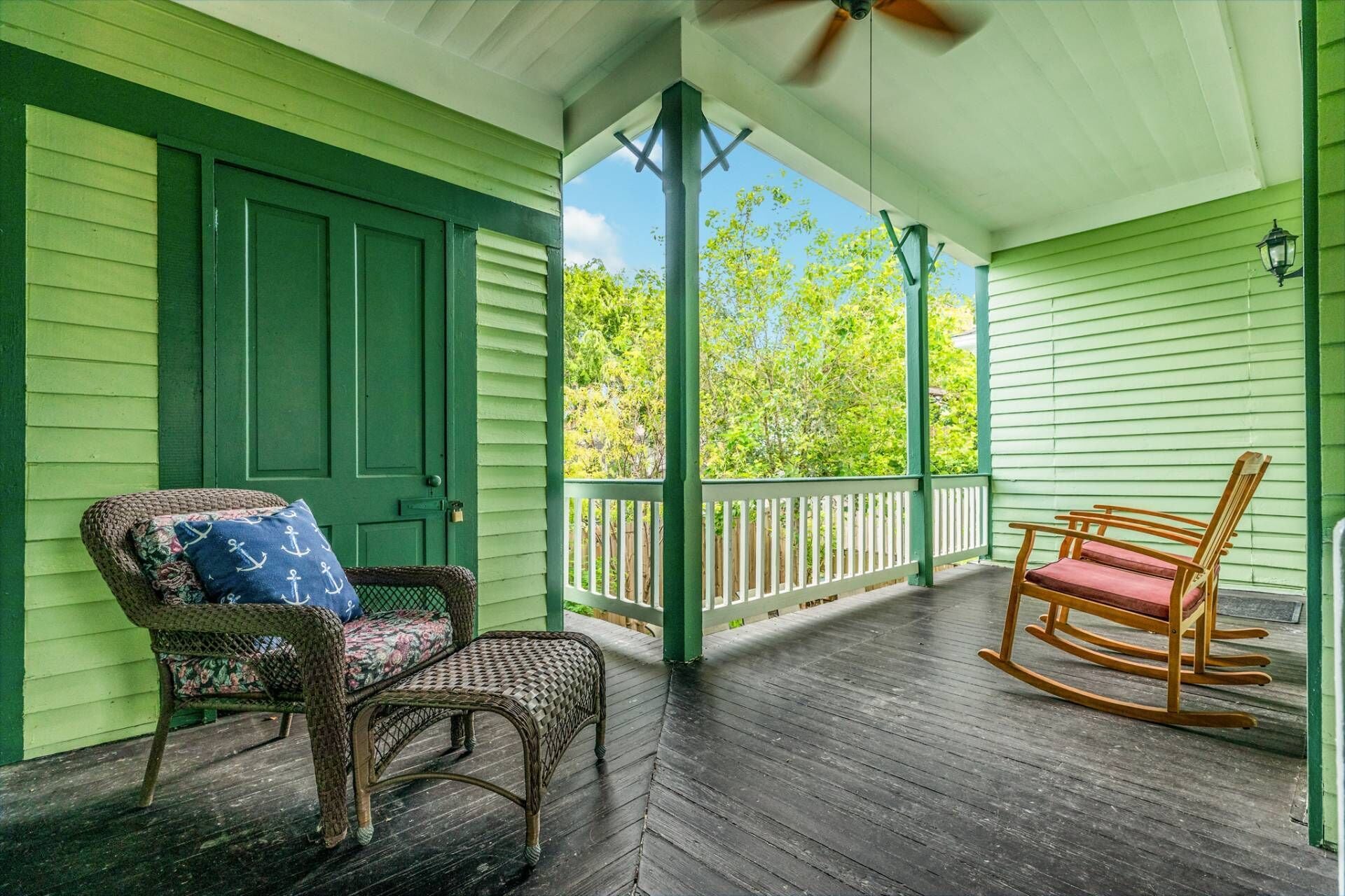 A porch with wicker chairs and a rocking chair, a green door, and a white railing.